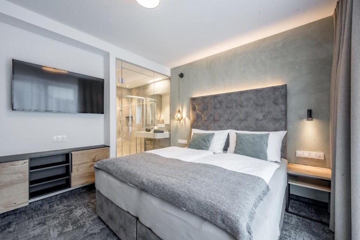 Ein modernes Hotelzimmer mit einem großen Bett, einem Flachbildfernseher, einem Spiegel und einem durch eine Glasabtrennung einsehbaren Badezimmer.