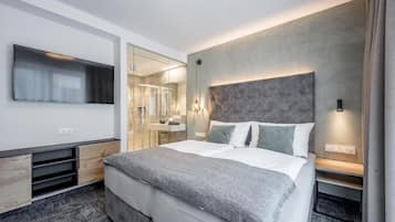 Habitación de hotel moderna con una cama grande, un televisor de pantalla plana, un espejo y un baño visible a través de un panel de vidrio.