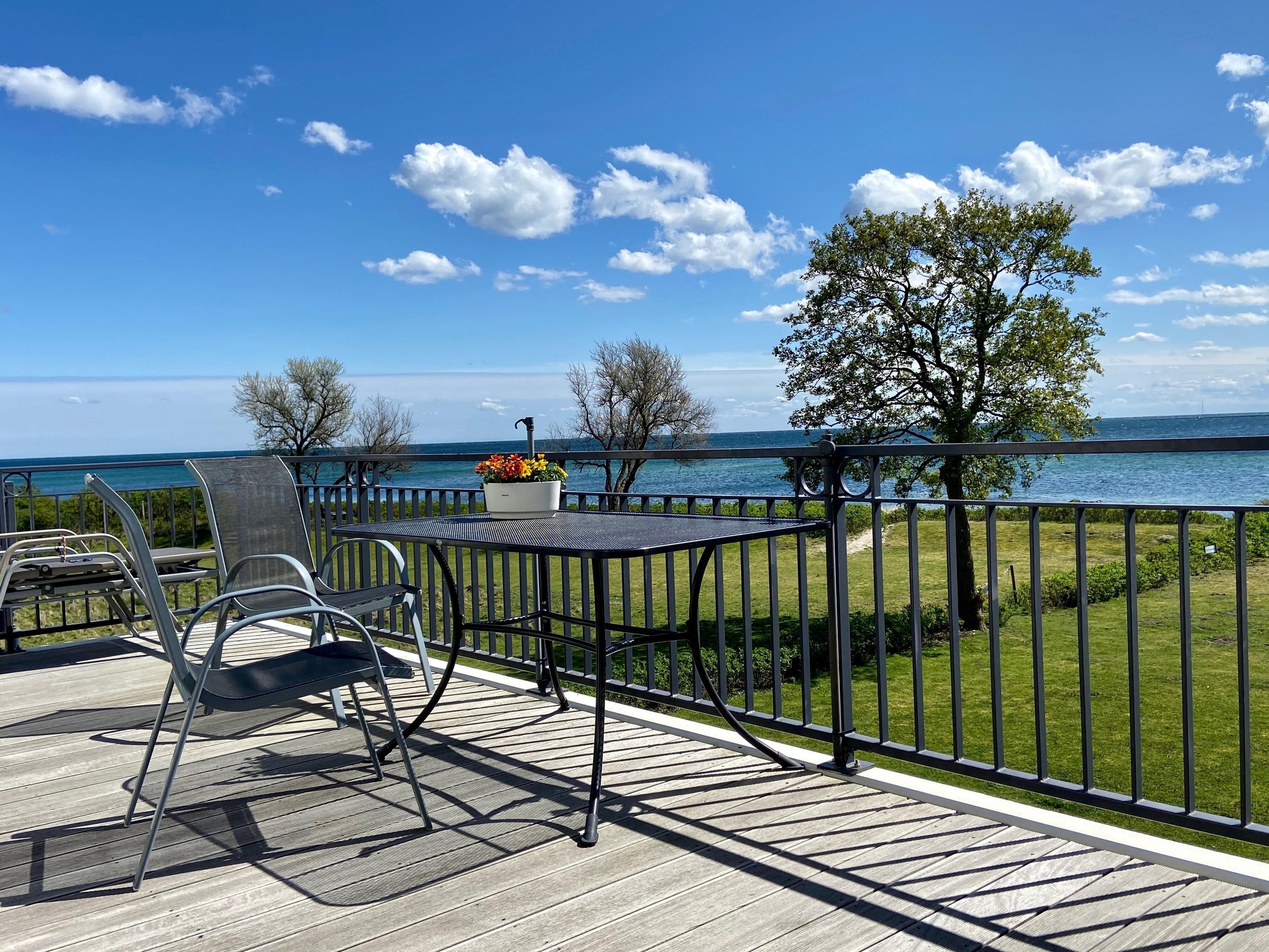A deck with a table and chairs overlooking a body of water, with a grassy area and trees in the background.