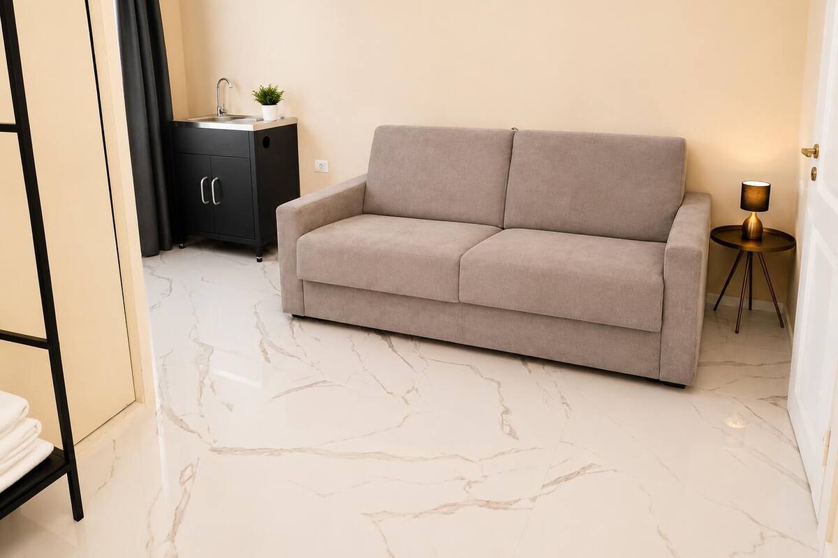 Ein modernes Wohnzimmer mit einem beigen Sofa, einer kleinen, schwarzen Ablage mit Waschbecken, einem runden Beistelltisch mit Lampe und einem Marmorboden.