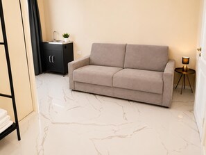 Un salon moderne avec un canapé beige, un petit meuble noir avec un évier, une table d’appoint ronde avec une lampe et un sol en marbre.