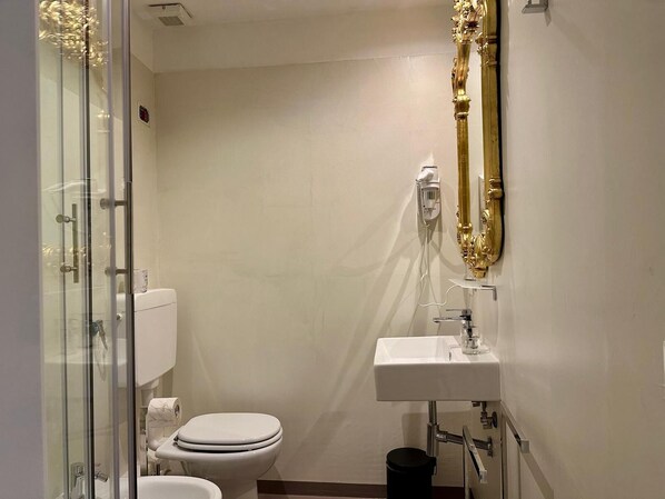 Egy kis fürdőszoba, amelyben WC, üvegfalas zuhanyzó és arany keretű tükör található.