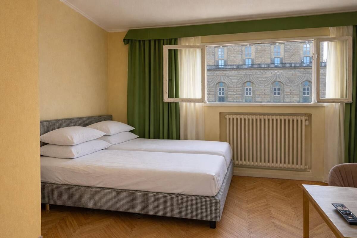 Ein Hotelzimmer mit Bett, Heizkörper, einem Fenster mit Vorhängen und Blick auf ein Gebäude.
