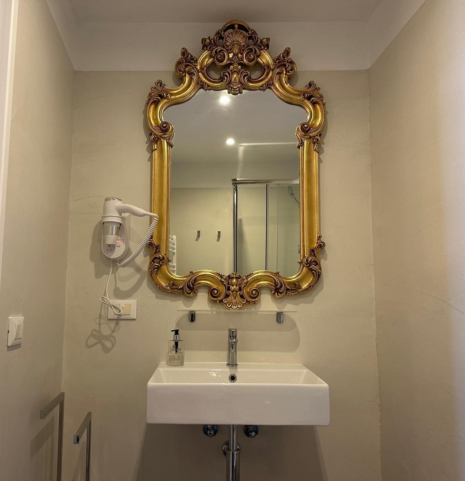 Ein Badezimmer mit einem goldfarbenen Spiegel, einem weißen Waschbecken und einem an der Wand befestigten Fön.