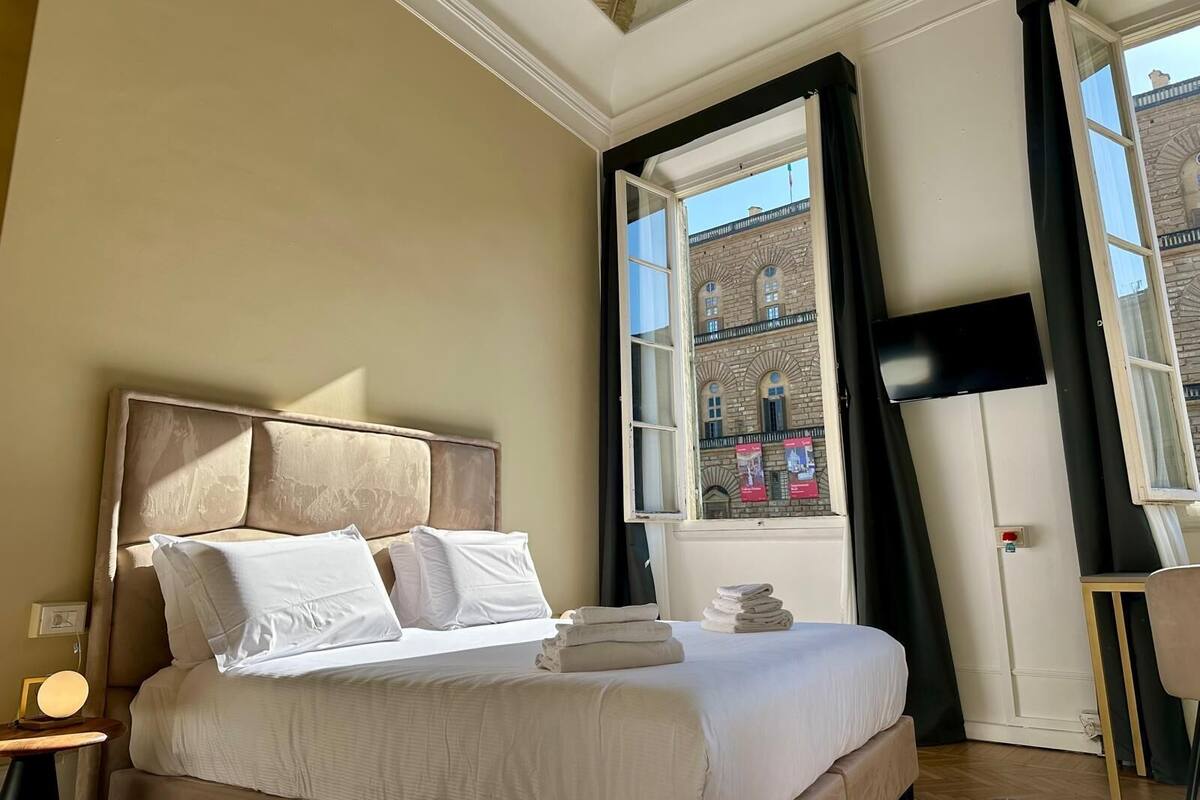 Ein Hotelzimmer mit einem großen Bett, einem Nachttisch, einem Schreibtisch und einem Fenster mit Blick auf ein Gebäude.