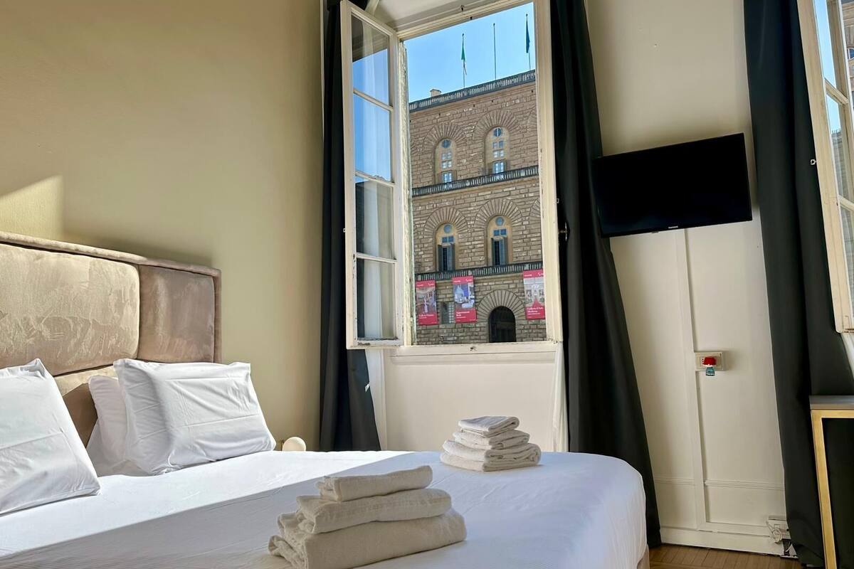 Ein Hotelzimmer mit Bett, Kissen, einem Stapel Handtücher, einem Fenster mit Blick auf ein Gebäude und einem an der Wand befestigten Flachbildfernseher.