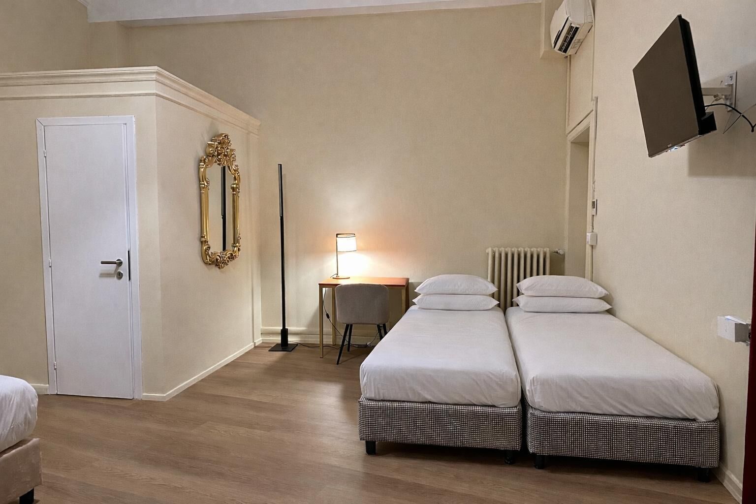 Ett hotellrum med två separata sängar, ett skrivbord med en stol, en lampa, en spegel och en TV som är monterad på väggen.
