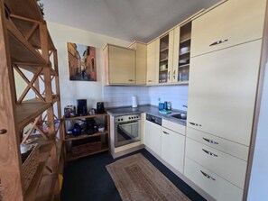 Une cuisine de taille réduite, équipée d’une cuisinière, d’un four et d’un évier. Un meuble-étagère en bois se trouve à gauche, et un tableau est accroché au-dessus des placards.