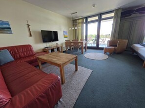 A furnished living room with a red sofa, a wooden coffee table, a flat-screen TV, and a balcony with outdoor seating.