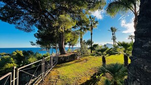 Un sentier côtier bordé de balustrades en pierre, entouré d’arbres et de plantes tropicales, qui mène à un point de vue sur la mer et une montagne lointaine.