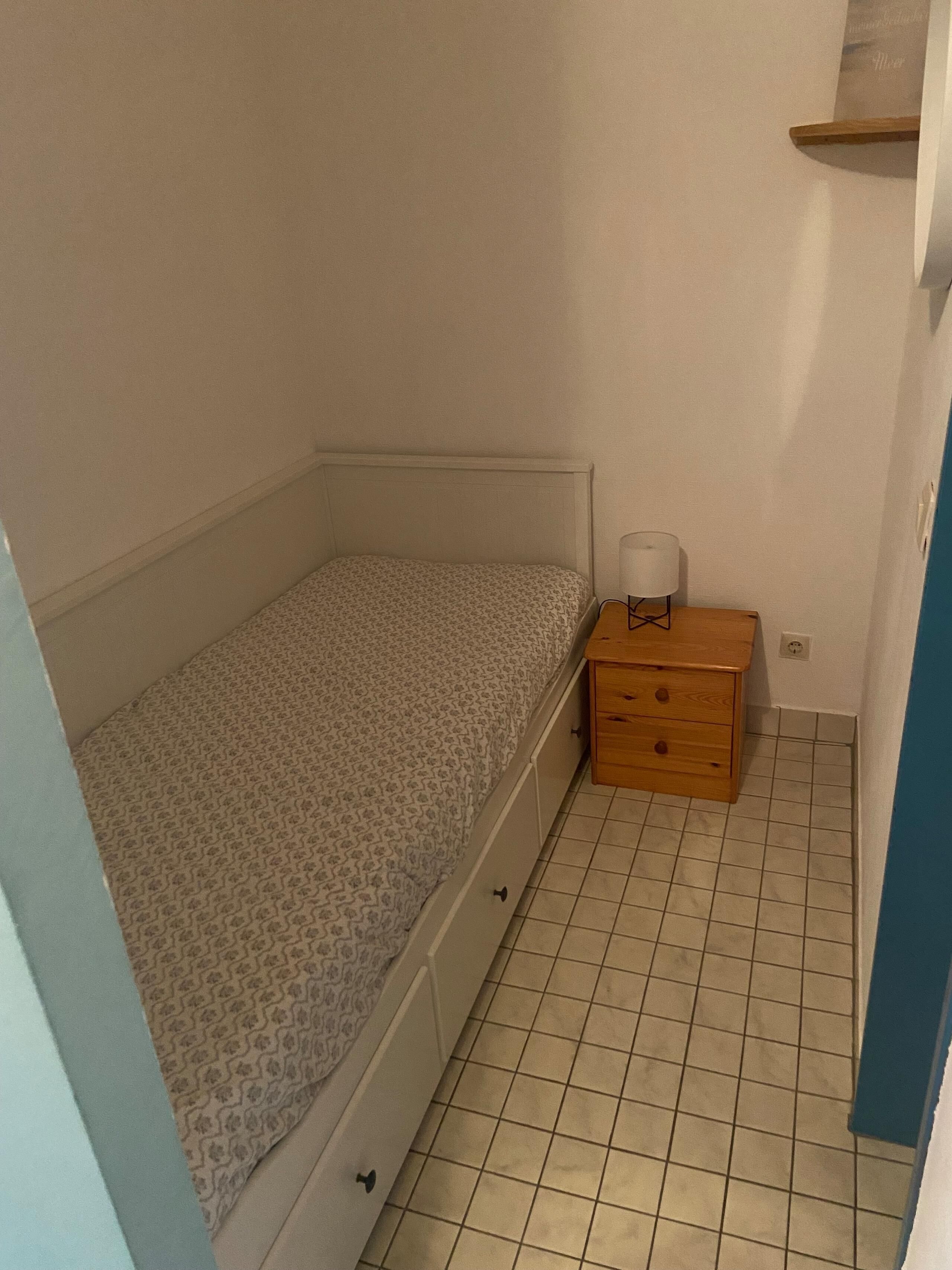 A small, single-bed room with a patterned bedspread, a wooden nightstand with drawers, and a tiled floor.