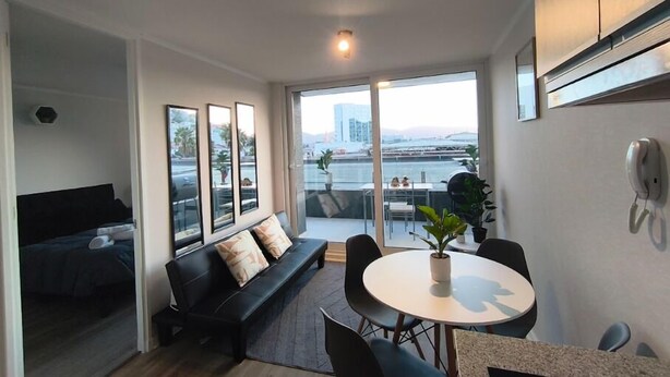 A modern living room with a sofa, a dining table, and a view of a cityscape through large windows. - Oceana Suites Edificio HA (Coquimbo)
