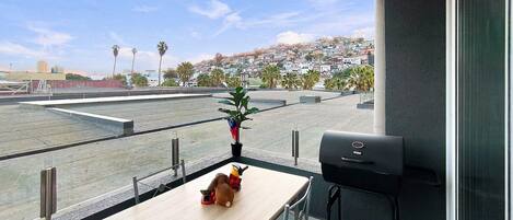 A balcony with a table, a grill, and a view of a cityscape.