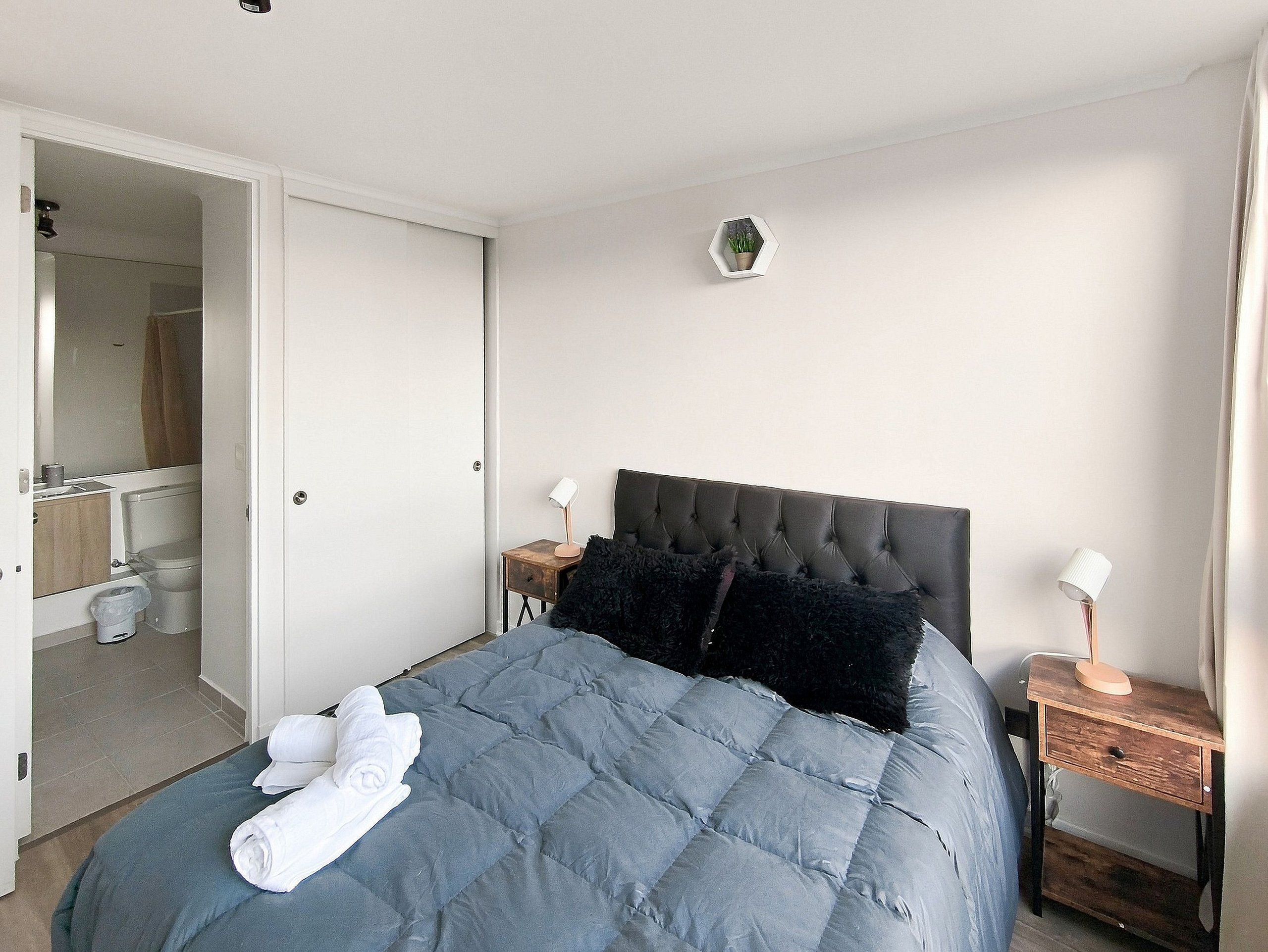 Een slaapkamer met een bed, nachtkastje, wandlamp, een potplant en een deur naar een badkamer.