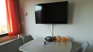 A dining area with a table, chairs, a flat-screen TV mounted on the wall, and a window with curtains. - Pure relaxation on the Baltic Sea: vacation apartment with sandy beach right on the doorstep! (Fehmarn)