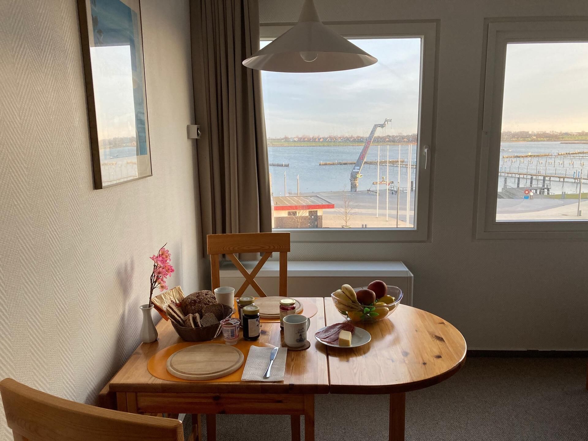 Uma área de refeições com uma mesa posta para dois, uma cadeira, uma janela com vista para um porto e uma luminária pendente.