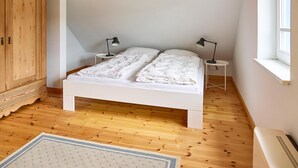 A bedroom with a wooden ceiling, white walls, a white bed, two bedside tables with lamps, a wooden wardrobe, and a window. - Relaxation and adventure at Deichhuus: your vacation paradise on the Baltic Sea! (Fehmarn)