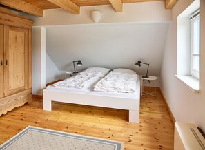 A bedroom with a wooden ceiling, white walls, a white bed, two bedside tables with lamps, a wooden wardrobe, and a window. - Relaxation and adventure at Deichhuus: your vacation paradise on the Baltic Sea! (Fehmarn)