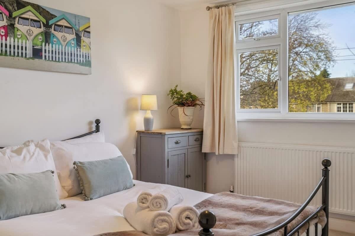 Une chambre à coucher avec un lit, des oreillers, une table de chevet avec une lampe, une plante en pot et une fenêtre avec des rideaux.