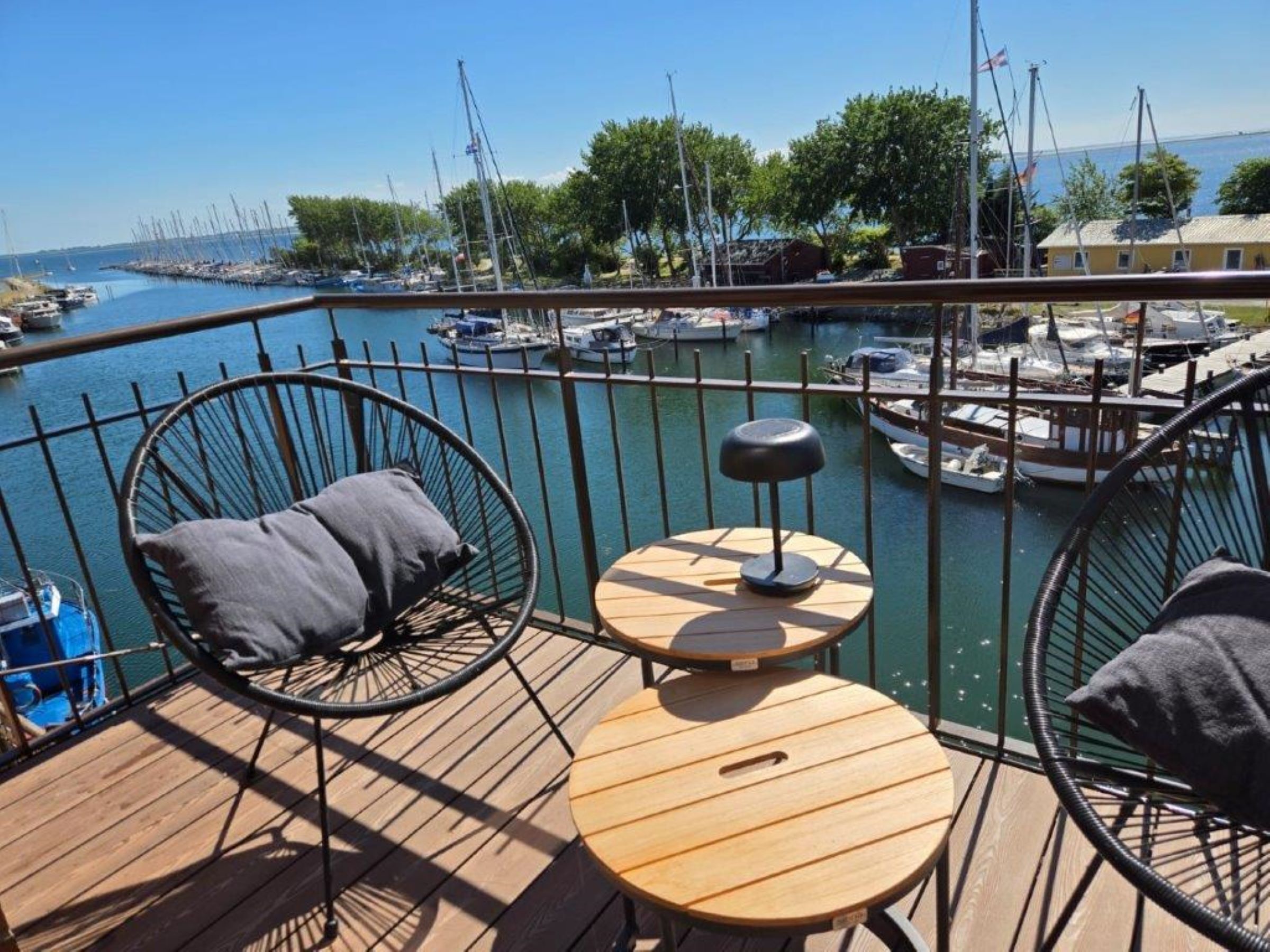 A wooden deck with two chairs and a small table, overlooking a marina with boats and a clear sky.