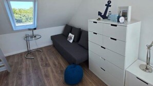 Habitación compacta con techo inclinado, una ventana pequeña, un sofá gris oscuro, un mueble de almacenamiento blanco y un sillón de peluche azul.