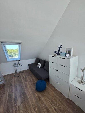 A compact room with a sloped ceiling, a small window, a dark gray sofa, a white storage unit, and a blue bean bag. - Toplicht Vacation Rental – Harbor View & Sauna in Orth (Schwanitz) with a Balcony and Lake View (Fehmarn OT Orth)
