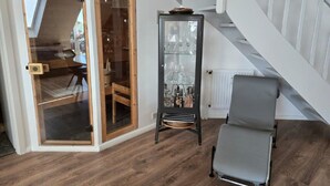 Una habitación con suelo de madera, una puerta de cristal que da a otra habitación, un armario negro con cristalería y una silla gris.