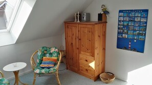 Une pièce avec un meuble en bois, une chaise en osier agrémentée d’un coussin coloré, une petite table ronde et une affiche représentant des phares.