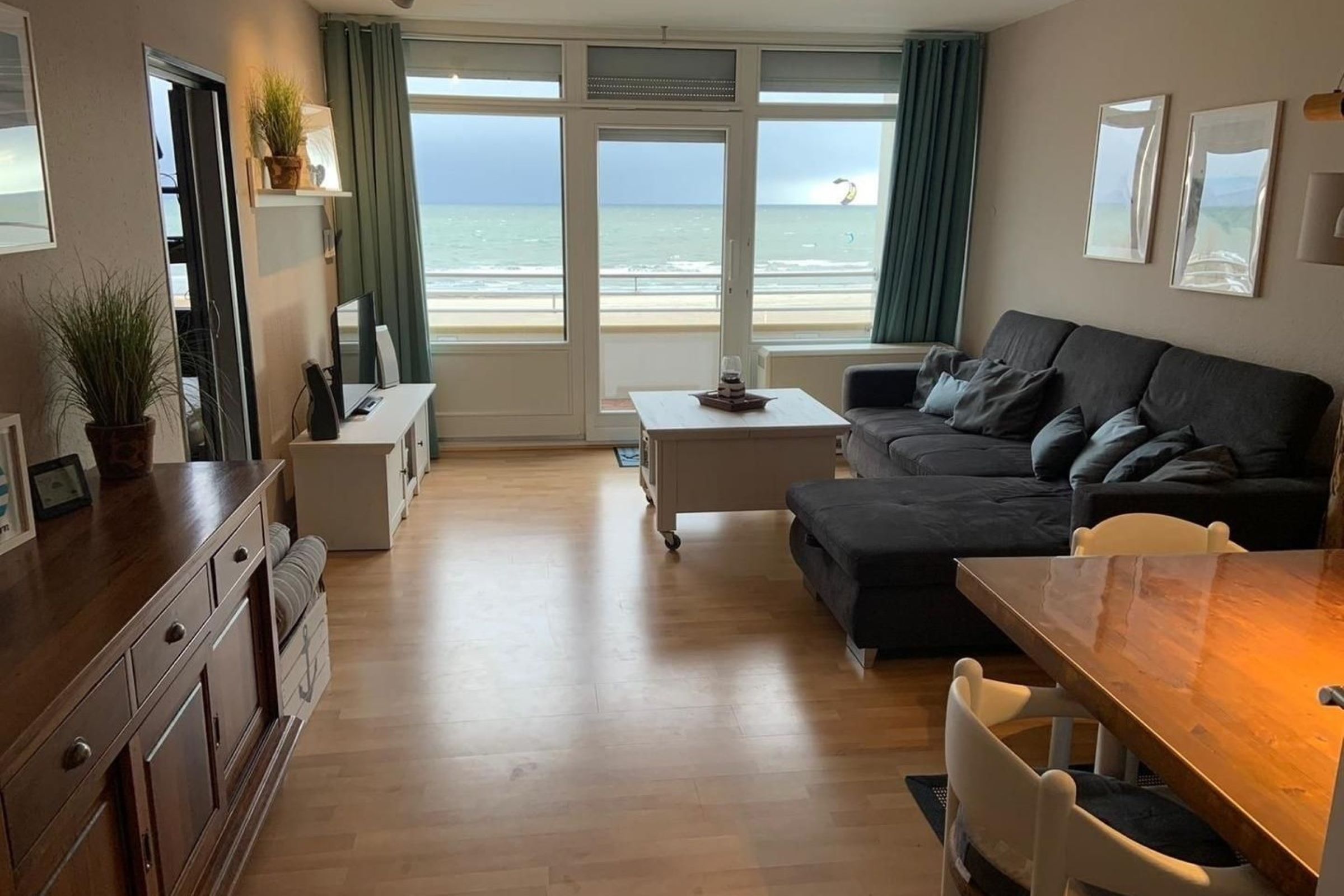 A modern living room with a sofa, coffee table, and a dining area with a wooden table and chairs. There is a view of the sea through large windows.