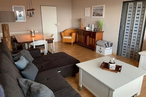 A living room with a sofa, a dining table, a cabinet, and a small white table with a glass and a tray. - Baltic Sea love: Your cozy vacation apartment with a fantastic sea view! (Fehmarn)