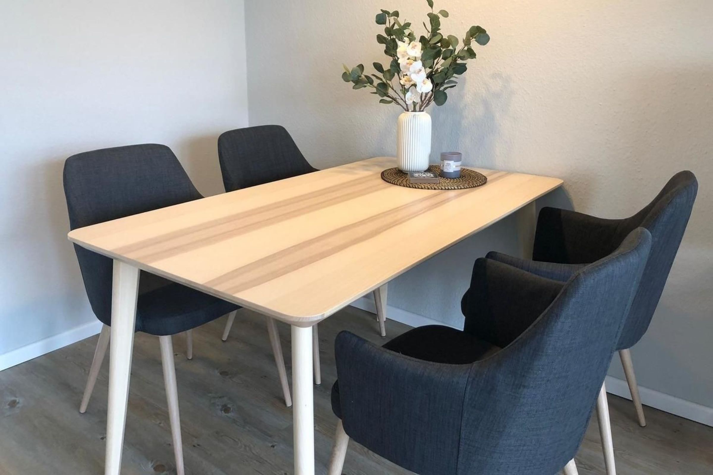 A dining table with four chairs, a vase with flowers, and a candle on a woven coaster.