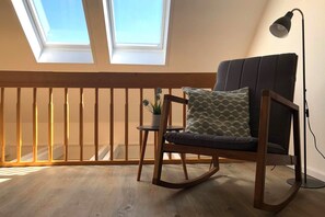 Une pièce avec un fauteuil à bascule en bois, une petite table avec une plante et un lampadaire. Il y a deux lucarnes au-dessus des fenêtres.