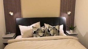 Ein ordentlich bezogenes Bett mit schwarzem Kopfteil, zwei Nachttische mit Lampen und eine Topfpflanze auf einem der Beistelltische.