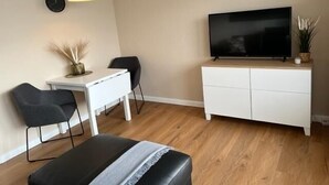 Ein modernes Wohnzimmer mit Flachbildfernseher, einem weißen Schrank, einem Schreibtisch, zwei Stühlen und einer Bank.
