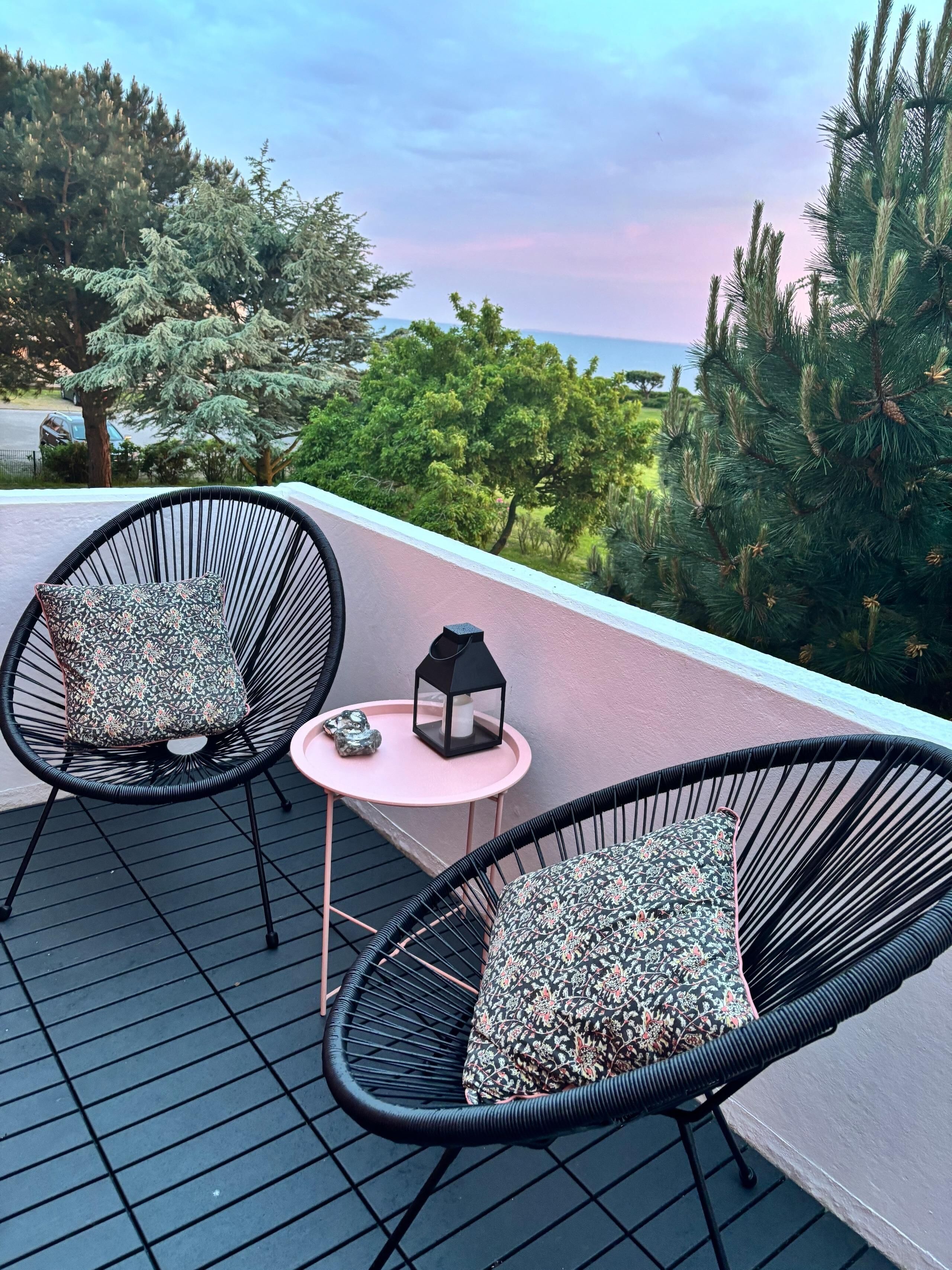Un balcon avec deux fauteuils en osier noir, chacun agrémenté d’un coussin à motifs, et une petite table rose sur laquelle est posée une lanterne.