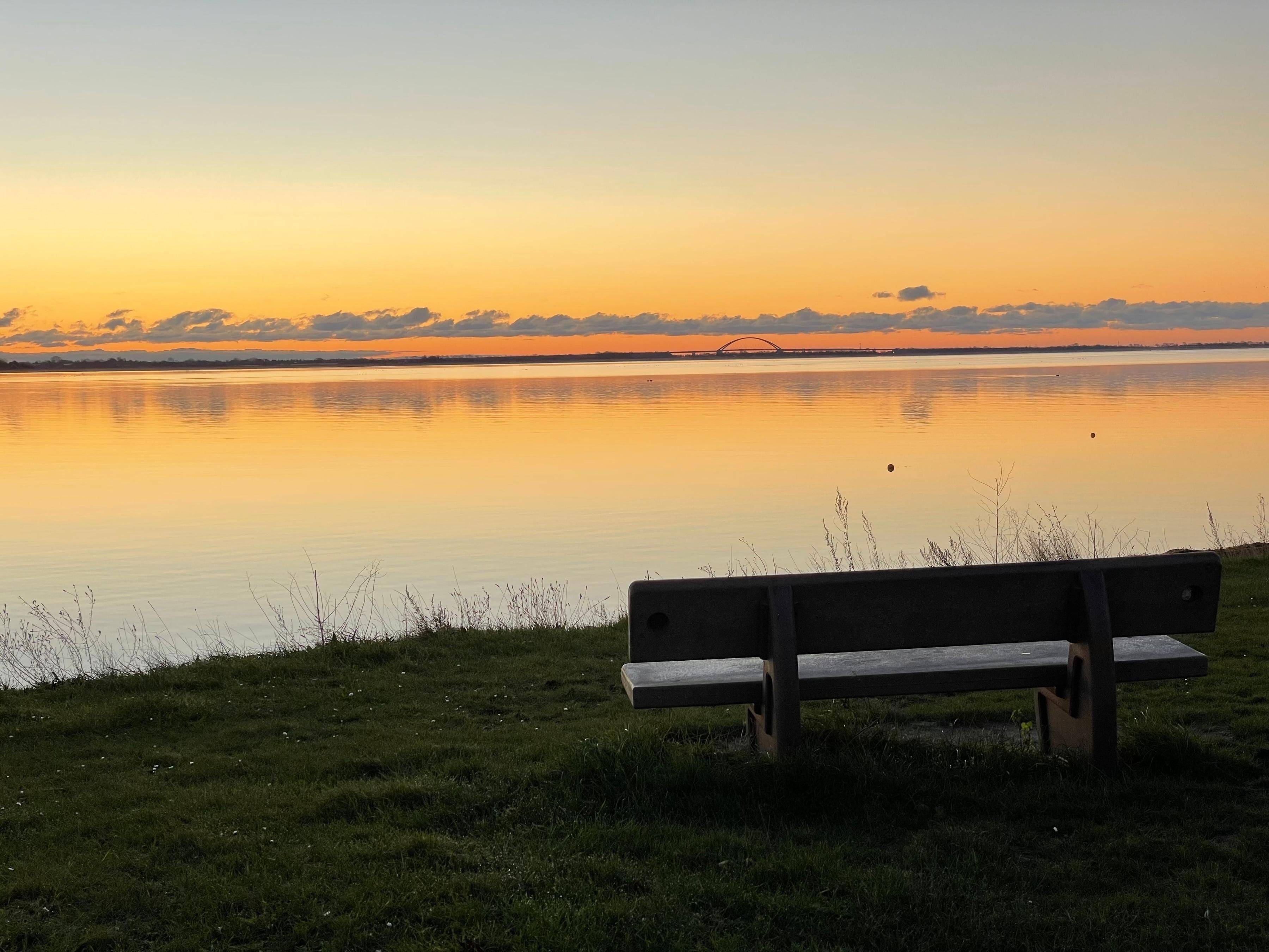 En bänk vid en stilla sjö i solnedgången, med en bro i fjärran.