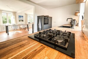 Et moderne køkken med en central ø, et spiseområde med bord og stole, samt udsigt til grønne omgivelser.