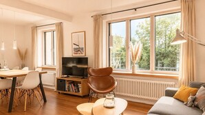 Ein modernes Wohnzimmer mit einer Couch, einem Esstisch mit Stühlen, einem Fernseher und einem großen Fenster mit Vorhängen.