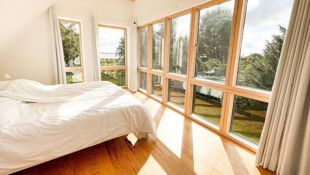 Ein Schlafzimmer mit einem großen Bett, Holzboden und mehreren Fenstern mit Blick auf Grünflächen und einen See.