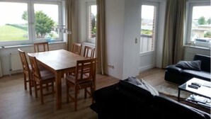 A room with a dining table and chairs, a black sofa, and a window with a view of a green landscape. - Vacation apartment with sea view on Fehmarn - Seeblick Marienleuchte (Fehmarn)