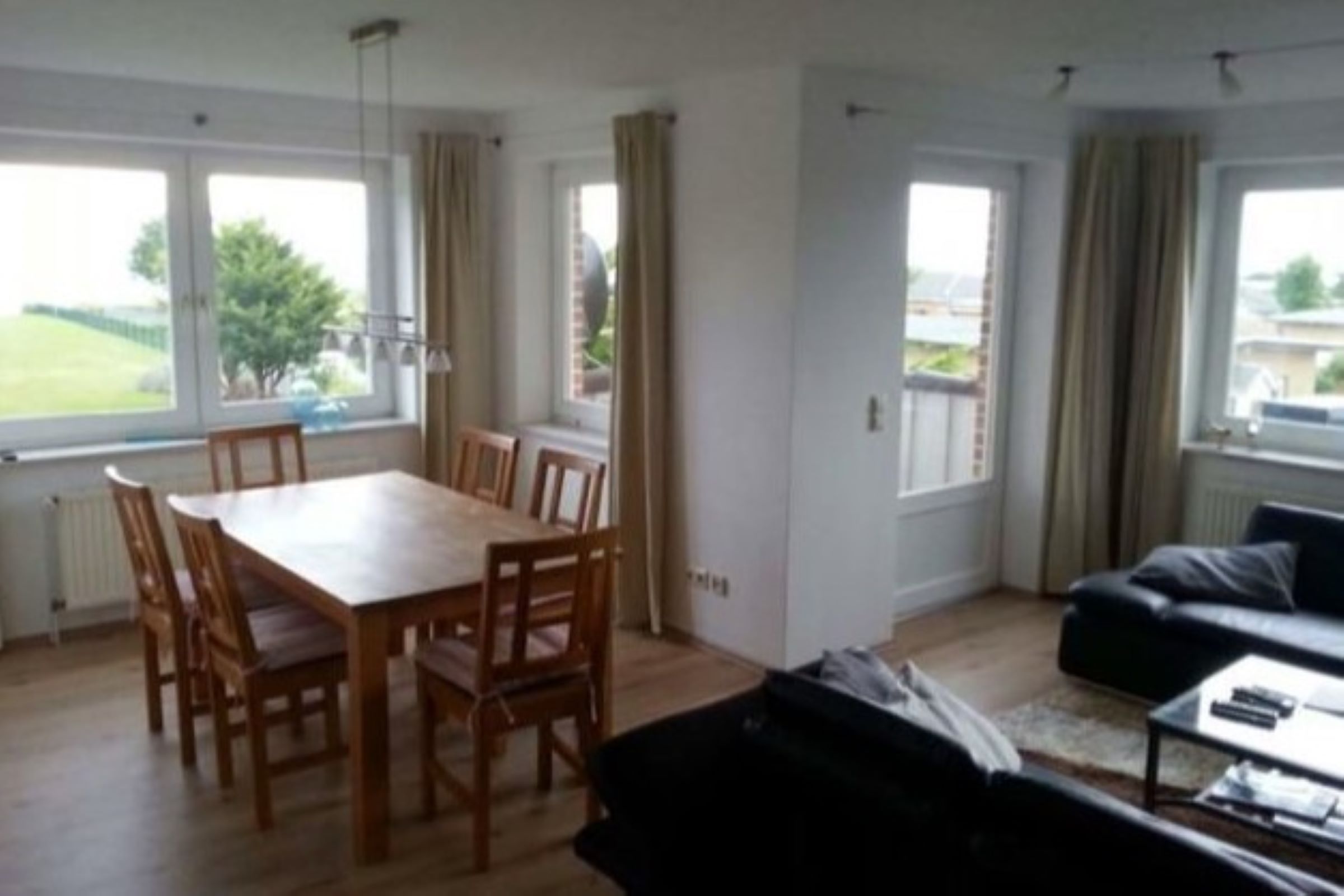 Ein Wohnzimmer mit Esstisch und Stühlen, einer Couch und einem Couchtisch. Große Fenster mit Vorhängen.
