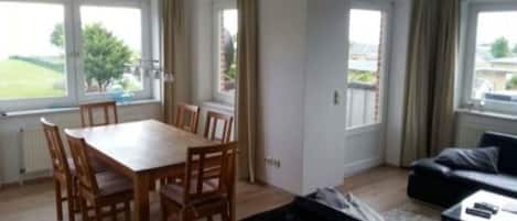 Ein Wohnzimmer mit Esstisch und Stühlen, einer Couch und einem Couchtisch. Große Fenster mit Vorhängen.