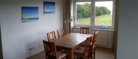 A dining area with a wooden table and chairs, a window with a view of greenery, and two paintings on the wall.