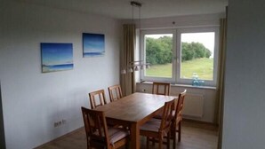 A dining area with a wooden table and chairs, a view of greenery outside, and two paintings on the wall. - Vacation apartment with sea view on Fehmarn - Seeblick Marienleuchte (Fehmarn)