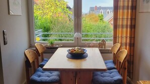 Un coin repas avec une table et des chaises en bois, un saladier rempli de fruits, et une vue sur les bâtiments et les arbres à travers de grandes fenêtres.