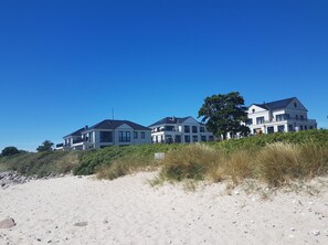 Ein Strandgrundstück mit mehreren Häusern, einem Sandstrand im Vordergrund und blauem, klarem Himmel.