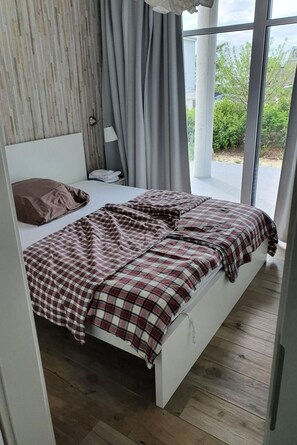 A bedroom with a bed, a nightstand, a lamp, and a view of the outdoors through large windows. - Only 40 m to the Baltic Sea - stylish vacation apartment on Fehmarnsund with terrace (Fehmarn)