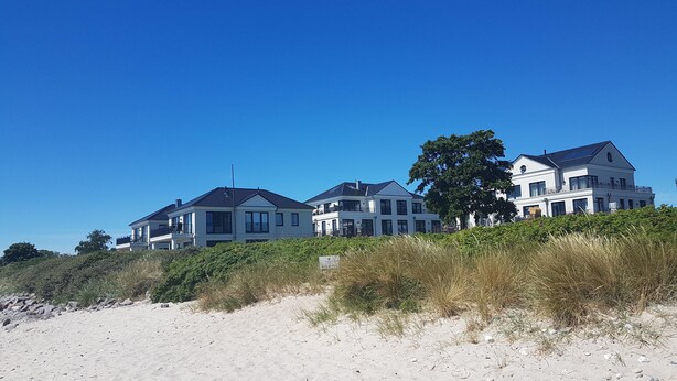 A beachfront property with multiple houses, a sandy beach in the foreground, and a clear blue sky. - Beach residence Vogelflug App. 10 - spacious & close to the beach (Fehmarn)