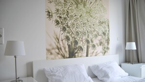 Ett hotellrum med en vit säng, två vita kuddar, ett sängbord med en lampa och en tavla som föreställer ett träd, upphängd på väggen.