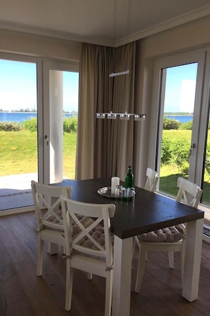 A dining area with a table and chairs, a view of the outdoors, and a curtain. - A relaxing beachfront vacation—a vacation apartment with a view of the sea! (Fehmarn)